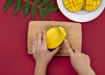 Draufsicht auf weibliche Hände, die Mango mit Messer auf Holzschneidebrett schneiden, mit geschnittener Mango auf weißem Teller und grünem Blatt auf rotem Hintergrund Marokko