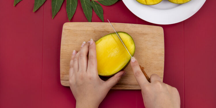 Draufsicht auf weibliche Hände, die Mango mit Messer auf Holzschneidebrett schneiden, mit geschnittener Mango auf weißem Teller und grünem Blatt auf rotem Hintergrund Marokko