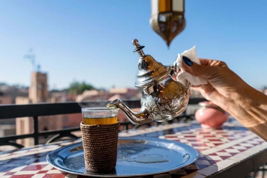Travelers enjoying traditional Moroccan tagine dinner on cozy rooftop restaurant during December winter getaway in Marrakech, with medina views and mountain backdrop at sunset