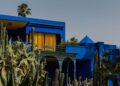 Bright blue modern house with yellow window curtains, set among tall cacti and palm trees against a clear sky.