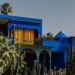 Bright blue modern house with yellow window curtains, set among tall cacti and palm trees against a clear sky.