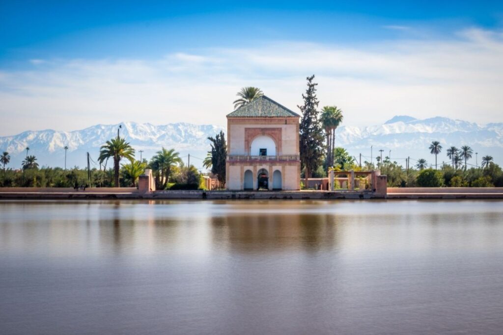 A small stone pavilion on the water with arches, palm trees, and distant snow-capped mountains under a blue sky.