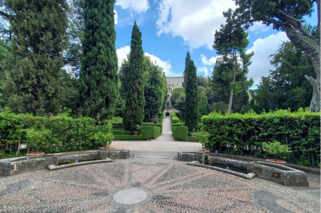 Formal garden with tall cypress trees, neatly trimmed hedges, and a stone-paved path toward a distant classical building.