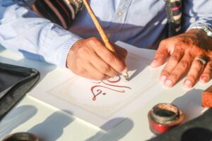 A Moroccan calligraphy artist holds a reed pen over paper covered in flowing Arabic script