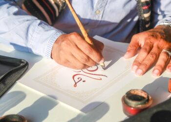 calligrapher-writing-arabic-script-unsplash - MAwebzine Een Marokkaanse kalligraaf houdt een rietpen boven papier bedekt met vloeiend Arabisch schrift