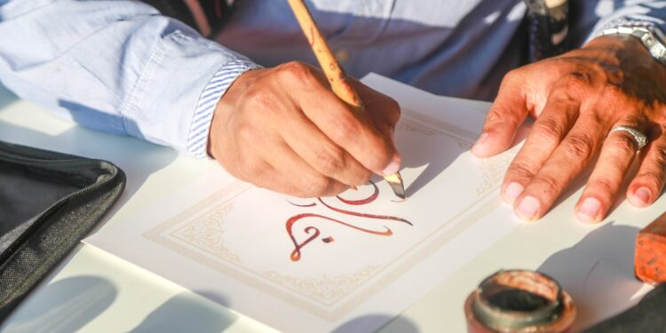 calligrapher-writing-arabic-script-unsplash - MAwebzine Een Marokkaanse kalligraaf houdt een rietpen boven papier bedekt met vloeiend Arabisch schrift