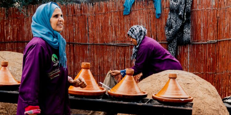 moroccan-woman-serving-tagine-pexels - MAwebzine Een Marokkaanse vrouw tilt het deksel van een aarden Marokkaanse tajine op aan een familietafel in een traditionele riadkeuken.