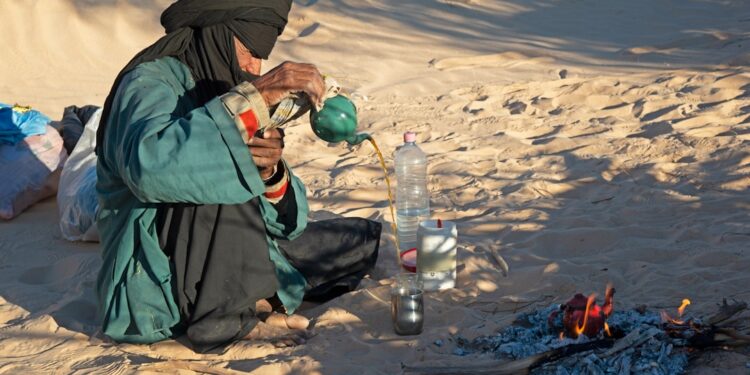 woman-pouring-moroccan-mint-tea-unsplash - MAwebzine Een vrouw die het traditionele hoge-giet-ritueel van Marokkaanse dranken uitvoert, die atay in kleine glazen serveert in een zonnige binnenplaats