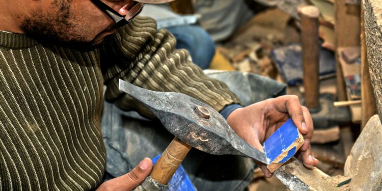 zellige tegeltegelbakker Marokko - MAwebzine Close-up van een ambachtsman die een blad op een steen scherpt, gekleed in een gestreepte trui en muts, met houten gereedschapsbehandelingen ergens in de buurt.